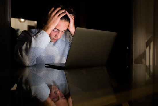 A picture of a person holding their head at night with an open laptop