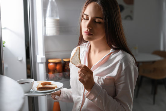 A person with their eyes closed, eating in front of their fridge