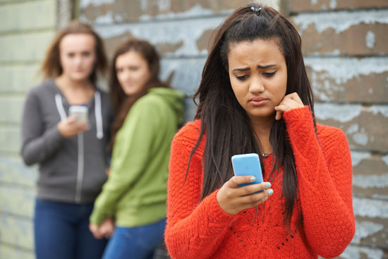 A young person frowning at their phone while two other teenagers look at them from behind