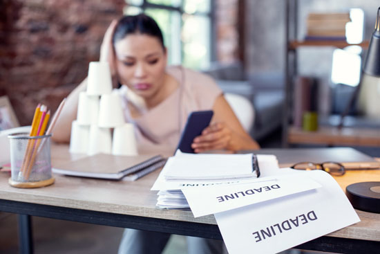 A person looking at their phone while a pile of papers titled 'Deadline' falls off their desk